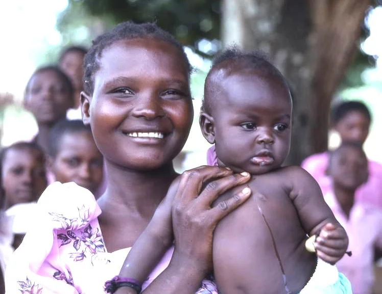 Photo of smiling Afro-Caribbean woman holding child level with her face, facing camera. In the backgound are other women looking on standing by a tree, out of focus