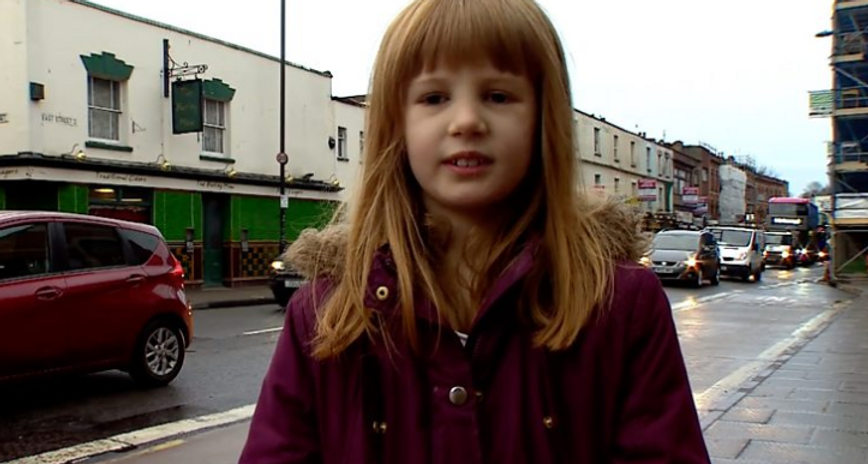 Photo of a red-headed girl wearing a duffel coat standing by a town street full of cars.