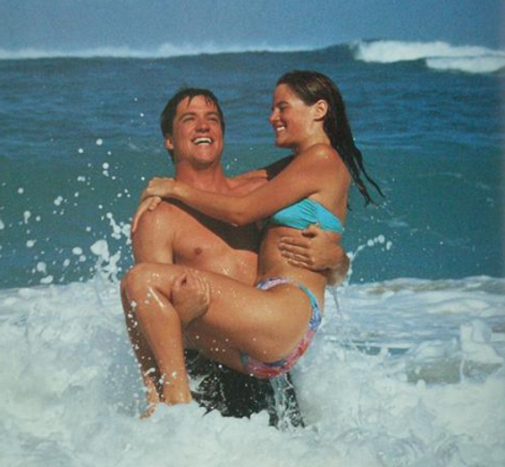 Photo of young couple in the sea. The man is standing in the surf, looking up and smiling while a smiling woman, possibly with eyes closed, is sitting in his arms being carried by him above the water. Their hair is wet, they wear swimsuits. Behind them a distant wave crests.