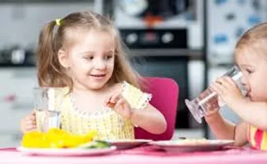 Two young children at kitchen table, eating and drinking. The girl, bunches in her hair, slightly older, looks at the boy (perhaps her younger brother) and smiles.