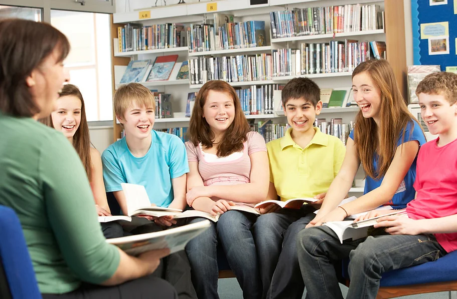 Photo of a group of smiling teenagers in a school library. A teacher, with open book, looking at them and laughing. The teens wear pastel colours, jeans and seem to be enjoying themselves.