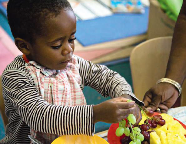 Photo of young Afro-Caribbean child sitting at a table, taking grapes off a bunch on a plate of fruit 