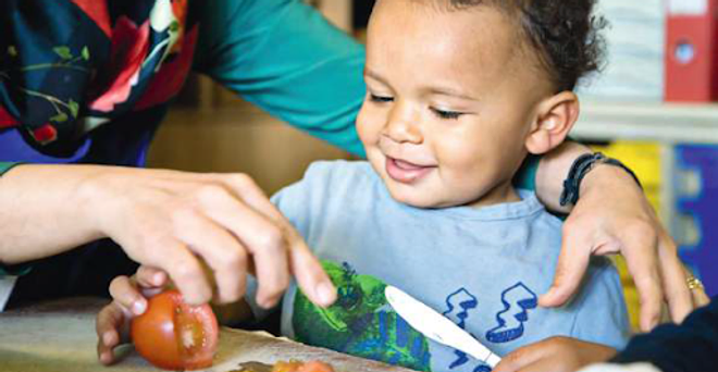 Photo of infant Afro-Caribbean child holding a knife and a sliced tomato, smiling. They are helped by Caucacian female arms, face off camera