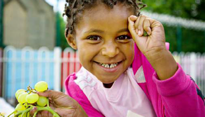 Photo of a young, smiling Afro-Caribbean child facing the camera, wearing pink and holding a bunch of grapes in one hand. With her other hand she touches her forehead.