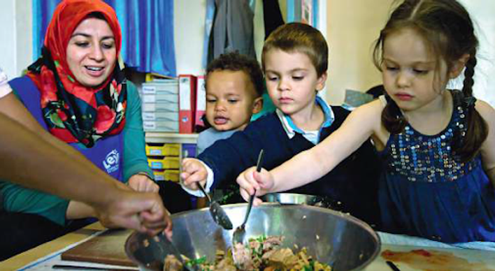 Photo of children of various ages cooking while being overseen by a woman in a hajib